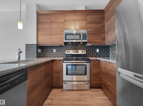 Kitchen featuring wood-finish cabinetry, stainless steel appliances, dark subway tile backsplash, light-toned countertops, and wood-finish flooring - #415 2588 Anderson Wy Sw, Edmonton, AB 