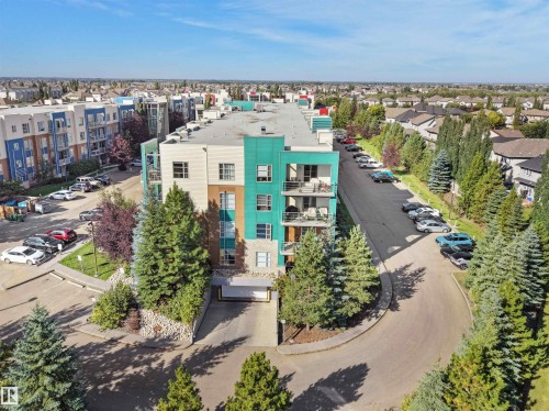 Modern multi-story building featuring balconies, an integrated parking garage, surrounding green spaces, and a paved circular driveway - #415 2588 Anderson Wy Sw, Edmonton, AB 