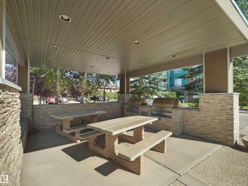 Covered outdoor entertaining area featuring two built-in grills, stone-clad walls, two concrete picnic tables, and recessed ceiling lighting - #415 2588 Anderson Wy Sw, Edmonton, AB 