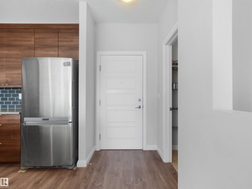 Stainless steel refrigerator, wood-finish flooring, dark wood-finish cabinetry, subway tile backsplash, and a modern paneled door - #415 2588 Anderson Wy Sw, Edmonton, AB 