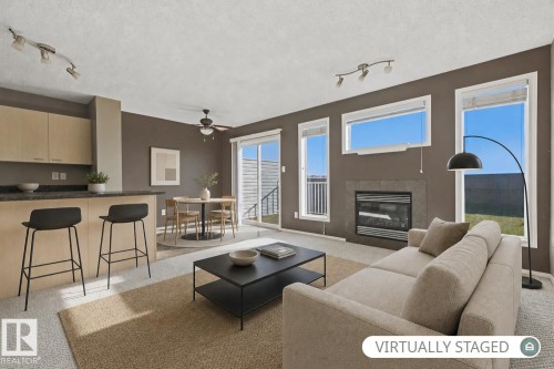 Open-concept living space featuring a gas fireplace with stone-finish surround, wood-finish kitchen cabinetry, breakfast bar, sliding glass door, and adjustable track lighting - 9047 Scott Crescent Nw, Edmonton, AB 