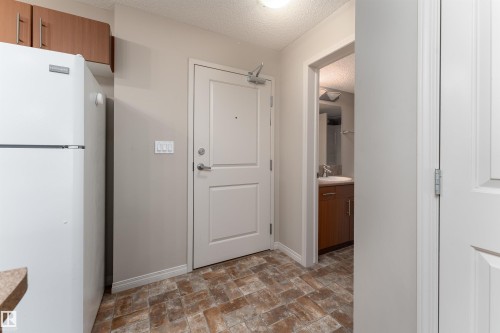White paneled door with brushed nickel hardware, textured flooring, and a white refrigerator with upper cabinetry - #202 105 Ambleside Dr Sw, Edmonton, AB 