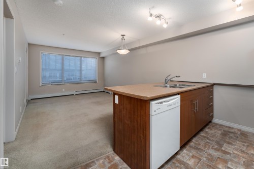 Open-concept living space featuring a central island with an integrated sink, wood-finish cabinetry, and a built-in dishwasher - #202 105 Ambleside Dr Sw, Edmonton, AB 