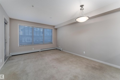 Neutral-toned carpeting, large window with horizontal blinds, overhead semi-flush mount light fixture, and baseboard heating units - #202 105 Ambleside Dr Sw, Edmonton, AB 