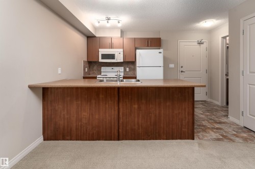 Kitchen area featuring wood-finish cabinetry, a stainless steel sink, and white appliances - #202 105 Ambleside Dr Sw, Edmonton, AB 