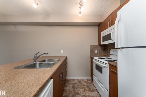 Kitchen featuring warm wood-finish cabinetry, solid surface countertops, a double basin stainless steel sink, and a full suite of white appliances, including a refrigerator, range, and microwave - #202 105 Ambleside Dr Sw, Edmonton, AB 