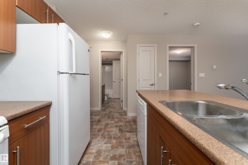 Kitchen with wood-finish cabinetry, light speckled countertops, and a double basin stainless steel sink - #202 105 Ambleside Dr Sw, Edmonton, AB 