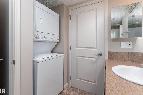 White stackable washer and dryer, vanity with integrated sink, patterned countertop, and a tiled backsplash - #202 105 Ambleside Dr Sw, Edmonton, AB 