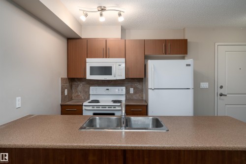 Kitchen featuring a dual basin stainless steel sink, wood-finish cabinetry, and a white appliance package - #202 105 Ambleside Dr Sw, Edmonton, AB 