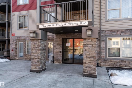 Stone facade entry with double glass doors, concrete walkway, exterior lighting fixtures, and prominent building address - #403 105 Ambleside Dr Sw, Edmonton, AB 