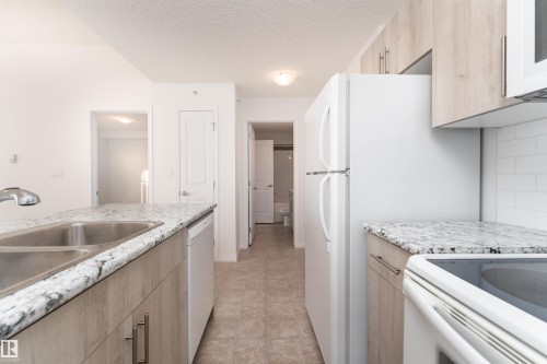 Kitchen featuring light wood-finish cabinetry, light-toned countertops, a double basin stainless steel sink, and white appliances - #403 105 Ambleside Dr Sw, Edmonton, AB 