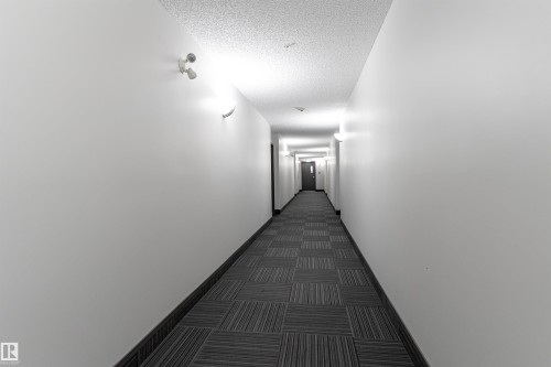 Long hallway with textured carpet flooring, sconce lighting, and a fire alarm - #403 105 Ambleside Dr Sw, Edmonton, AB 