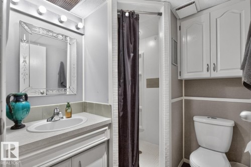 White vanity with an integrated sink and chrome faucet, decorative mirror with sconce lighting, white shower stall with a dark privacy curtain, white built-in storage cabinetry, and a white toilet - 5502 48 Avenue, Beaumont, AB 