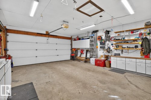 Interior garage featuring a white overhead door, concrete flooring, overhead fluorescent lighting, and built-in white cabinetry with wood-finish countertops - 5502 48 Avenue, Beaumont, AB 