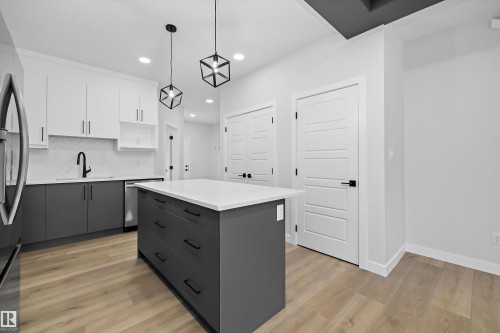 Kitchen featuring two-tone cabinetry, central island with stone-finish countertop, geometric pendant lighting, herringbone-patterned backsplash, and wood-finish flooring - Edmonton, AB 