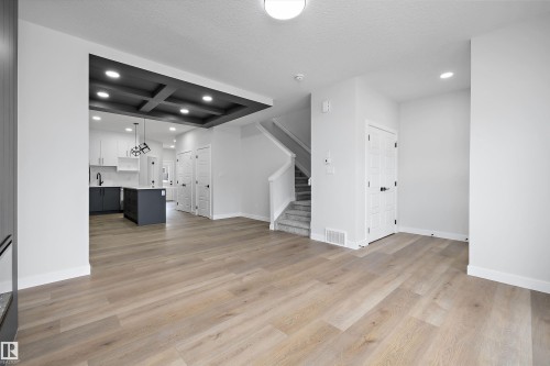 Open-concept interior featuring wood-finish flooring, a coffered ceiling with recessed lighting, a kitchen island with contrasting cabinetry, and a staircase with neutral carpeting - Edmonton, AB 