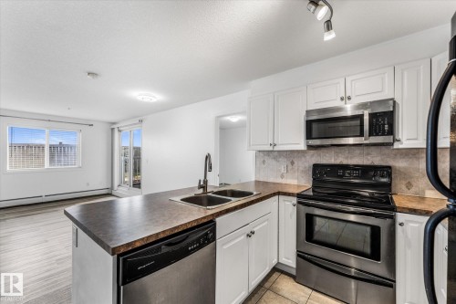 Kitchen featuring white cabinetry, dark countertops, a double-basin stainless steel sink with a high-arch faucet, and a suite of stainless steel appliances - #109 4309 33 St, Stony Plain, AB 
