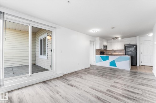 Open-concept interior featuring wood-finish flooring, a kitchen with white cabinetry, a peninsula, and glass sliding doors leading to a private patio - #109 4309 33 St, Stony Plain, AB 