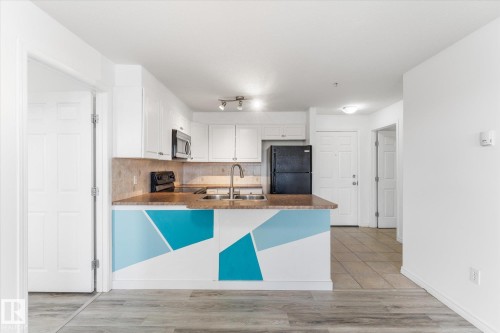 Kitchen featuring white cabinetry, solid-surface countertops, dual-basin stainless steel sink with high-arc faucet, and tiled backsplash - #109 4309 33 St, Stony Plain, AB 