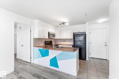Kitchen featuring white cabinetry, solid-surface countertops, dual-basin stainless steel sink with arch faucet, and wood-finish flooring - #109 4309 33 St, Stony Plain, AB 
