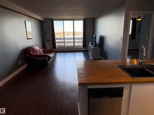 Open-concept living space featuring wood-finish flooring, floor-to-ceiling glass sliding doors, and a kitchen peninsula with a wood-style countertop, stainless steel dishwasher, and undermount sink - #1304 10160 116 St Nw, Edmonton, AB 