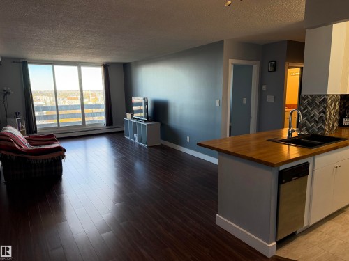 Open-concept interior featuring wood-finish flooring, floor-to-ceiling sliding glass doors, a kitchen island with a butcher block countertop, a matte black dual-basin sink, and a herringbone-patterned tile backsplash - #1304 10160 116 St Nw, Edmonton, AB 