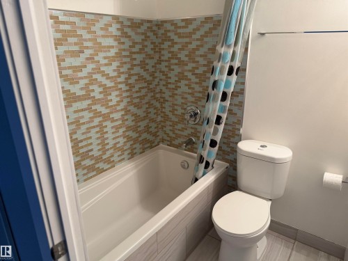 Bathtub enclosure featuring multi-colored mosaic tile wall detailing, light-toned rectangular tile surround, dual-flush toilet, and wall-mounted metal towel bar - #1304 10160 116 St Nw, Edmonton, AB 