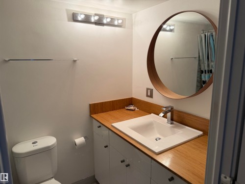 Bathroom featuring a wood-finish countertop, white integrated sink with a chrome single-handle faucet, white cabinetry, circular wood-frame mirror, and multi-bulb wall-mounted lighting - #1304 10160 116 St Nw, Edmonton, AB 