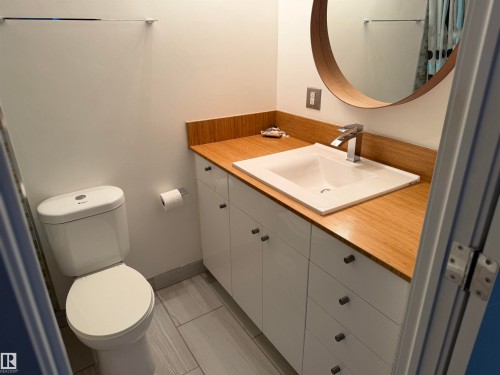 Bathroom vanity featuring a wood-finish countertop, white cabinetry with dark hardware, a rectangular drop-in sink, and a circular wall-mounted mirror - #1304 10160 116 St Nw, Edmonton, AB 
