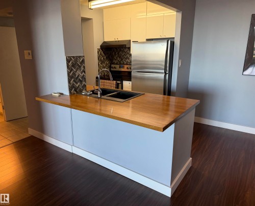 Kitchen featuring a peninsula with a wood-finish countertop, dual-basin undermount sink, herringbone-pattern tile backsplash, stainless steel appliances, and flat-panel cabinetry - #1304 10160 116 St Nw, Edmonton, AB 