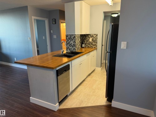 Kitchen featuring white cabinetry, wood-finish countertops, a black dual-basin sink, a herringbone pattern tile backsplash, and tile flooring - #1304 10160 116 St Nw, Edmonton, AB 