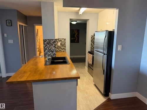 Kitchen featuring wood-finish countertop, dual-basin sink with arched faucet, herringbone-pattern tile backsplash, stainless steel refrigerator, and wood-finish flooring - #1304 10160 116 St Nw, Edmonton, AB 