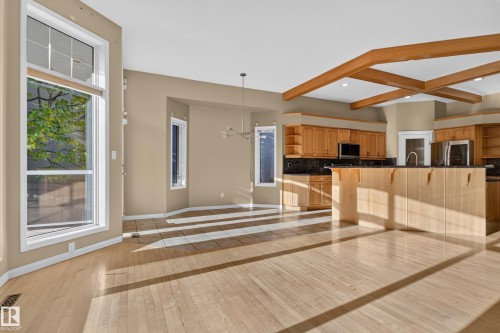 Open-concept kitchen and living area featuring exposed wood ceiling beams, wood-finish flooring, full-height windows, and a central kitchen island with built-in cabinetry - 876 Twin Brooks Close Nw, Edmonton, AB 
