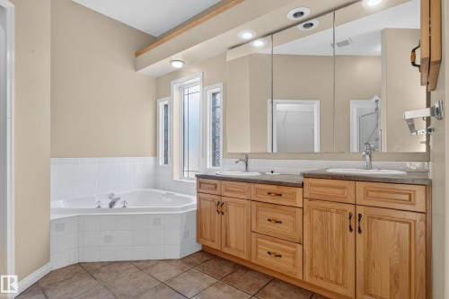 Bathroom featuring a dual-sink vanity with wood cabinetry and stone-finish countertops, large jetted soaking tub with tiled surround, and expansive wall-to-wall vanity mirror with integrated lighting - 876 Twin Brooks Close Nw, Edmonton, AB 
