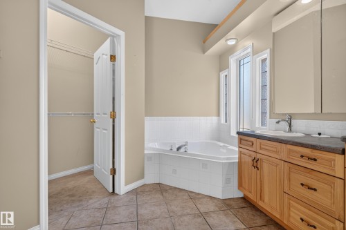 Bathroom featuring a corner soaking tub with white tile surround, wood cabinetry, stone-finish countertop, undermount sink, and tiled flooring - 876 Twin Brooks Close Nw, Edmonton, AB 