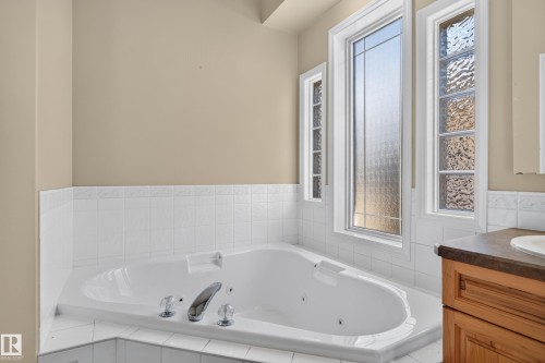 Corner jetted soaking tub featuring white tile surround, wood-finish cabinetry, and textured glass windows - 876 Twin Brooks Close Nw, Edmonton, AB 