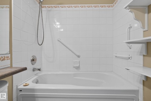 Full-size soaking tub featuring white tile surround, handheld shower fixture, chrome hardware, and white wall-mounted shelving - 876 Twin Brooks Close Nw, Edmonton, AB 