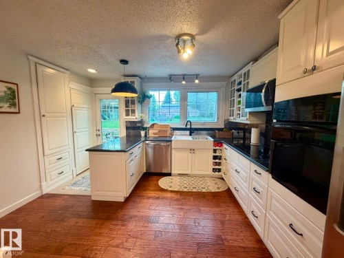 Kitchen featuring white cabinetry, dark countertops, a farmhouse sink, stainless steel appliances, and wood-finish flooring - 5 Gilmore Crescent, St. Albert, AB 