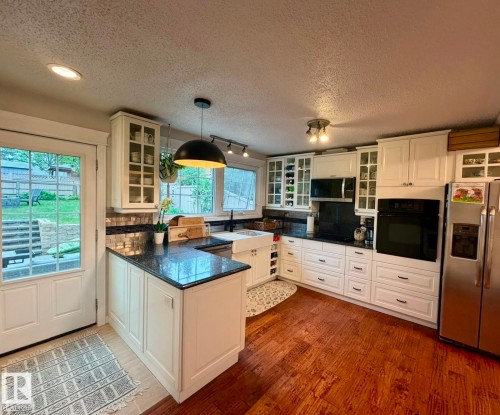 U-shaped kitchen with white cabinetry, dark stone countertops, wood-finish flooring, a white farmhouse sink, and stainless steel appliances - 5 Gilmore Crescent, St. Albert, AB 