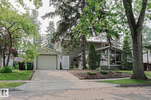 Two-story home featuring a single-car garage with an overhead door, a front-facing sunroom with expansive windows, and a landscaped front yard with mature trees - 5 Gilmore Crescent, St. Albert, AB 