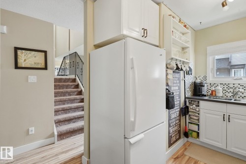 Wood-finish flooring, white cabinetry with dark hardware, checkered tile backsplash, window over sink, and carpeted stairs with a wrought iron railing - 5340 38A Avenue Nw, Edmonton, AB 