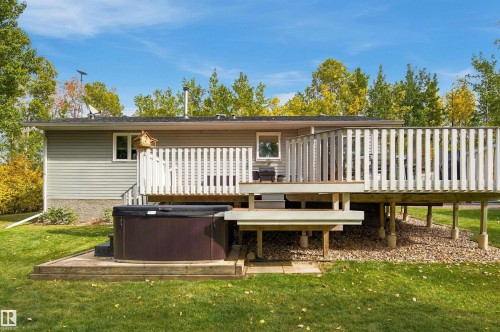 Expansive deck with white railings and built-in steps, featuring a spa with integrated steps, set against a backdrop of mature trees - 51064 Rge Rd 222, Rural Strathcona County, AB 