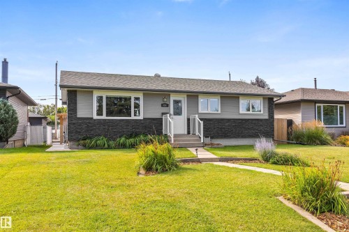 Single-story exterior featuring a grey lap siding and dark stone facade, white-trimmed windows, a paved concrete walkway, and landscaped garden beds - 6207 85 Avenue Nw, Edmonton, AB 