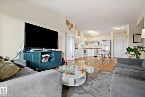 Open-concept living space with wood-finish flooring, white kitchen cabinetry, stainless steel refrigerator, and recessed lighting - #416 111 Edwards Dr Sw, Edmonton, AB 