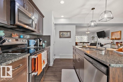 Kitchen featuring dark wood cabinetry, stone countertops, stainless steel appliances, a horizontal tile backsplash, and hanging pendant lighting - 6818 Evans Wynd Nw, Edmonton, AB 