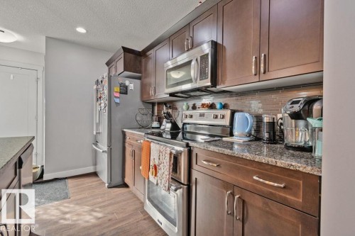 Kitchen featuring dark wood-finish cabinetry, stone-finish countertops, stainless steel appliances, and a linear tile backsplash - 6818 Evans Wynd Nw, Edmonton, AB 