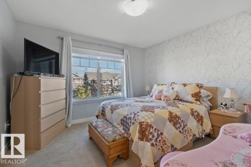 Neutral-toned bedroom featuring patterned wallpaper, carpeted flooring, and a sliding window with muntins - 6818 Evans Wynd Nw, Edmonton, AB 