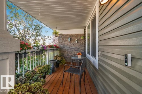 Covered porch featuring wood-finish decking, natural stone veneer accent wall, white metal railing, and horizontal vinyl siding - 6818 Evans Wynd Nw, Edmonton, AB 