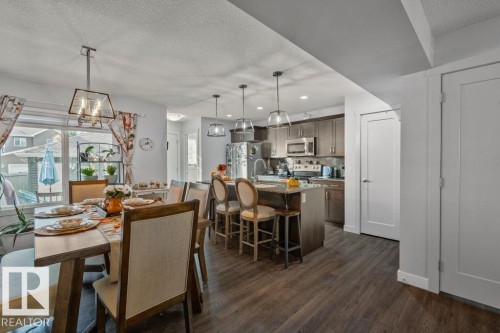 Kitchen and dining area featuring dark-tone cabinetry, a central island with breakfast bar seating, stainless steel appliances, pendant lighting, and wood-finish flooring - 6818 Evans Wynd Nw, Edmonton, AB 