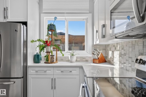 Kitchen featuring white shaker-style cabinetry, solid-surface countertops, a mosaic tile backsplash, stainless steel appliances, and a window with a roller shade - 798 Berg Loop, Leduc, AB 